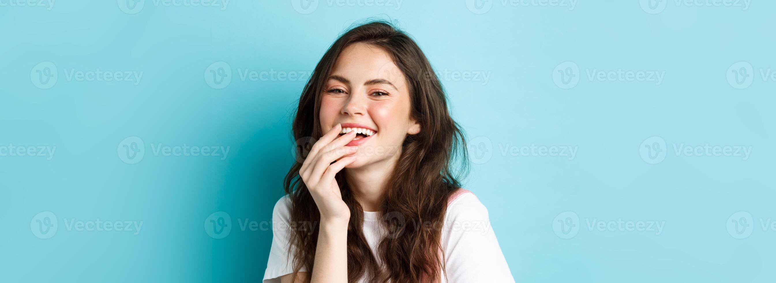 Close up of attractive young woman with stylish curly hairstyle and make up, laughing, covering her smile with hand and look at camera, standing over blue background photo