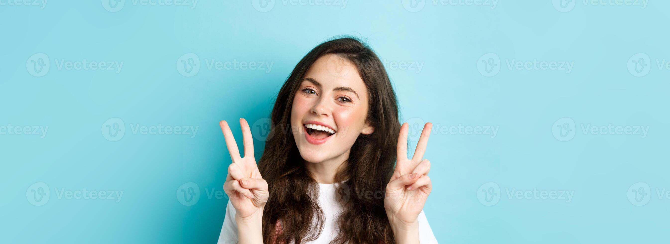 Young positive modern girl sending good vibes only, showing v-sign and smiling happy, standing against blue background photo