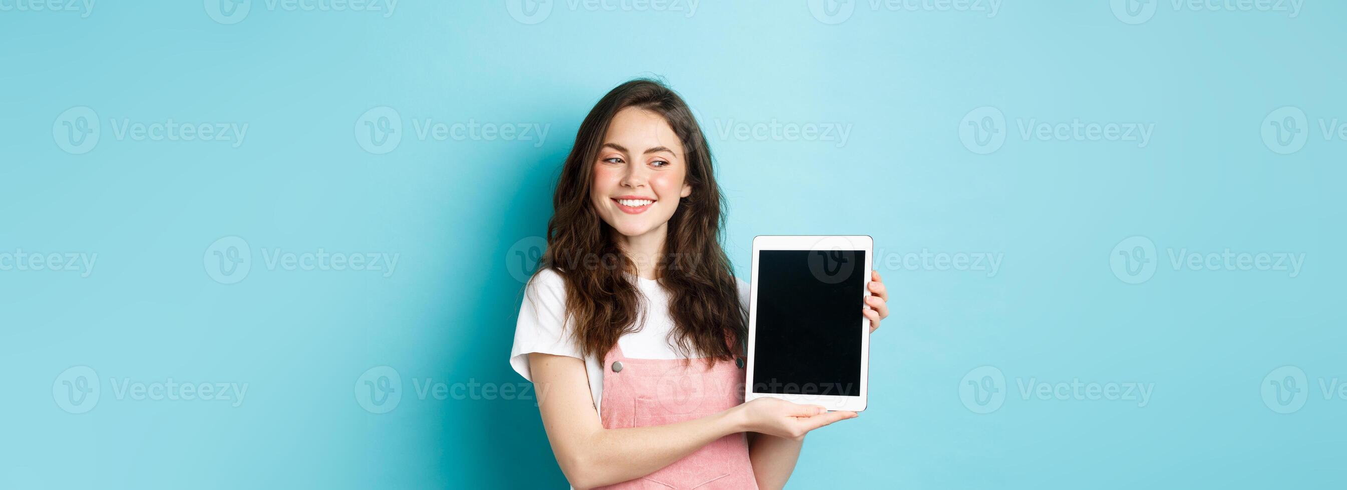 Image of happy young girl showing digital tablet screen and smiling proud, showing your logo on display, standing over blue background photo