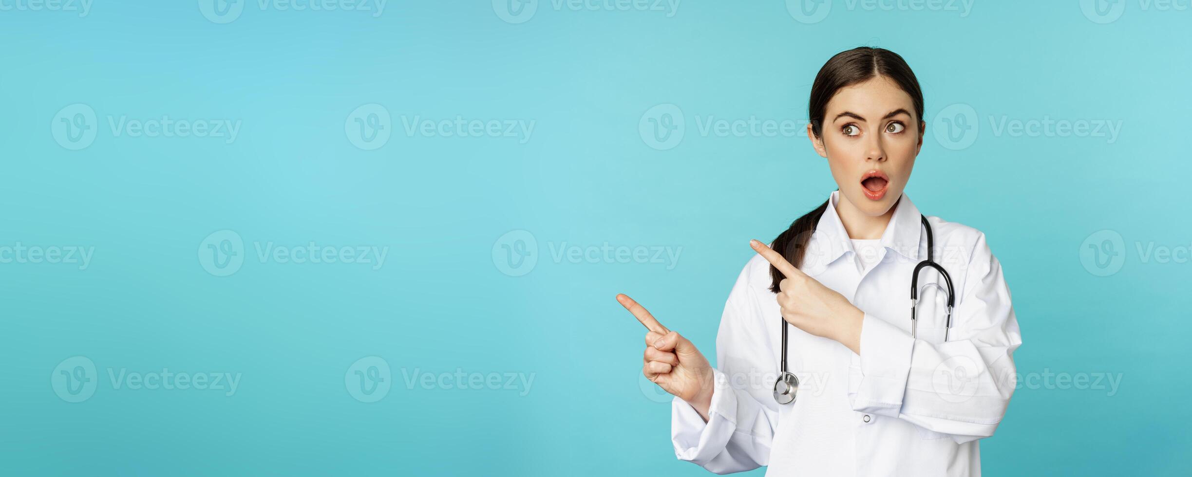 Surprised female doctor, physician with stethoscope, pointing and looking left with amazed, wow face, standing over torquoise background photo