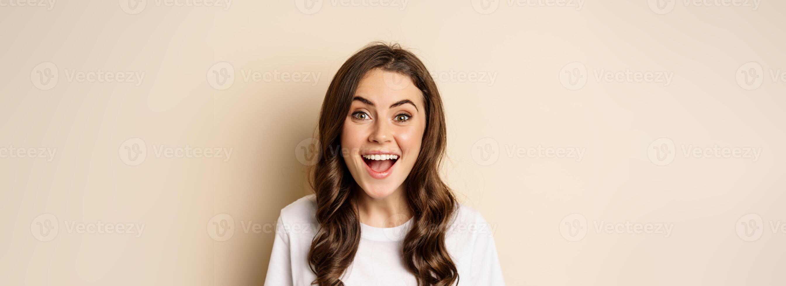 Close up portrait of surprised and excited brunette girl looking amazed, reacting impressed and excited, standing against beige background photo