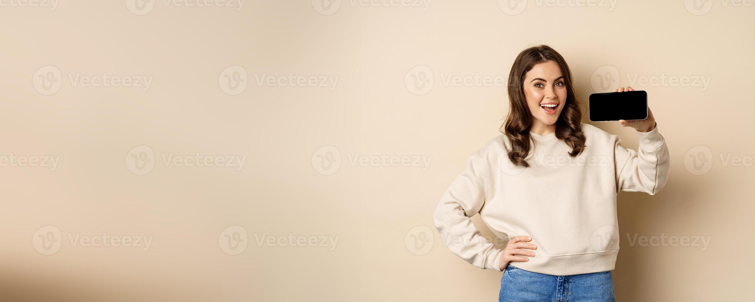 Smiling beautiful female model demonstrating horizontal smartphone screen, mobile application, standing over beige background photo