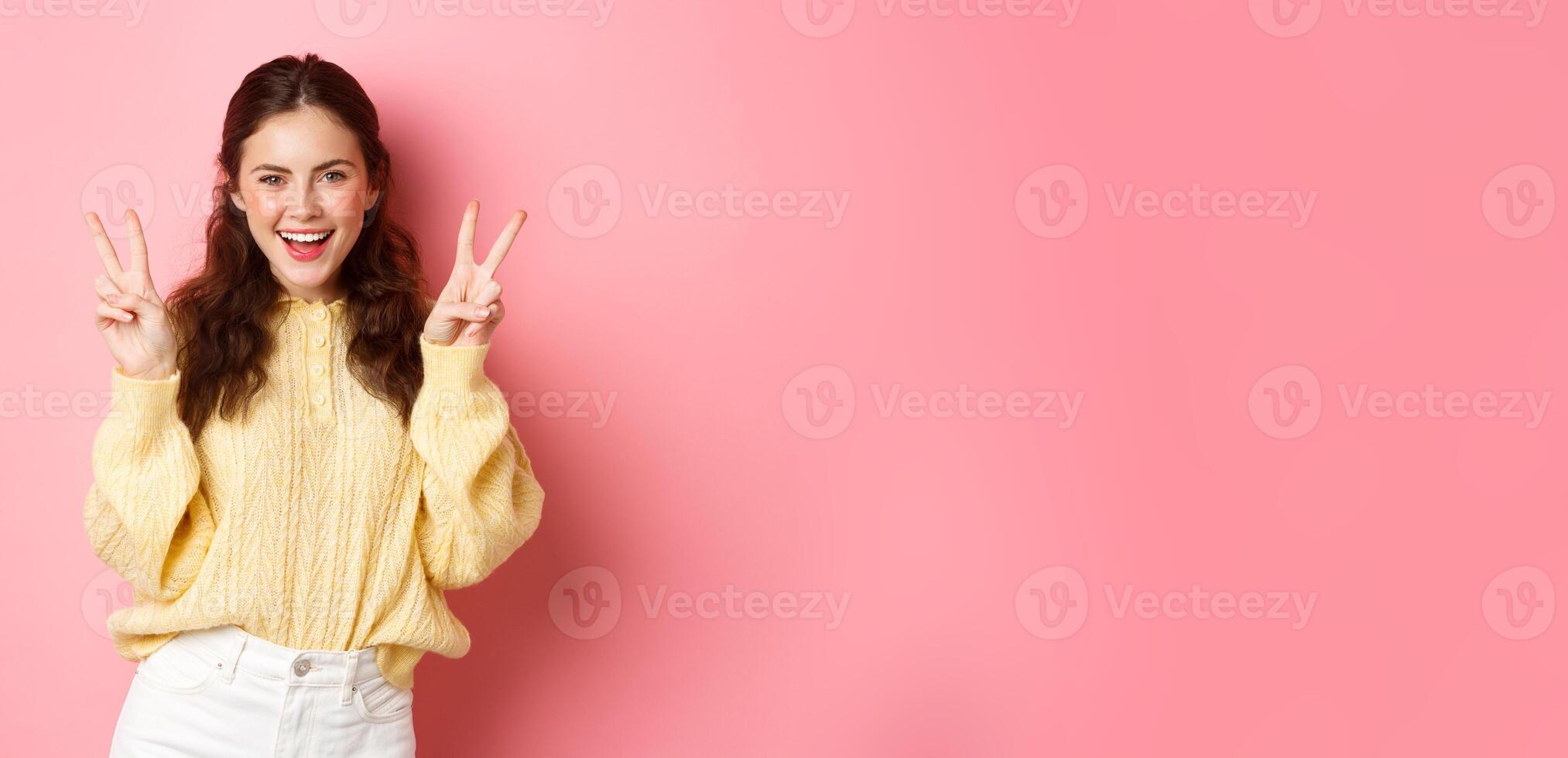 Portrait of attractive brunette female model, smiling and showing peace signs, making v-sign gesture, standing positive and happy against pink background photo