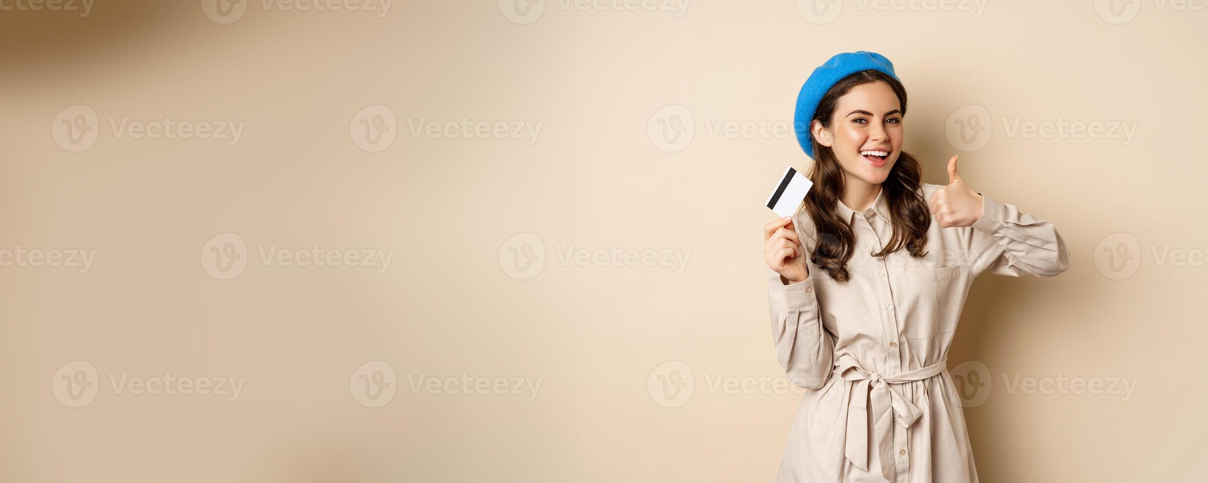 Concept of shopping and sale. Happy young woman in stylish clothes, showing credit card store discount and thumbs up, recommending, standing over beige background photo