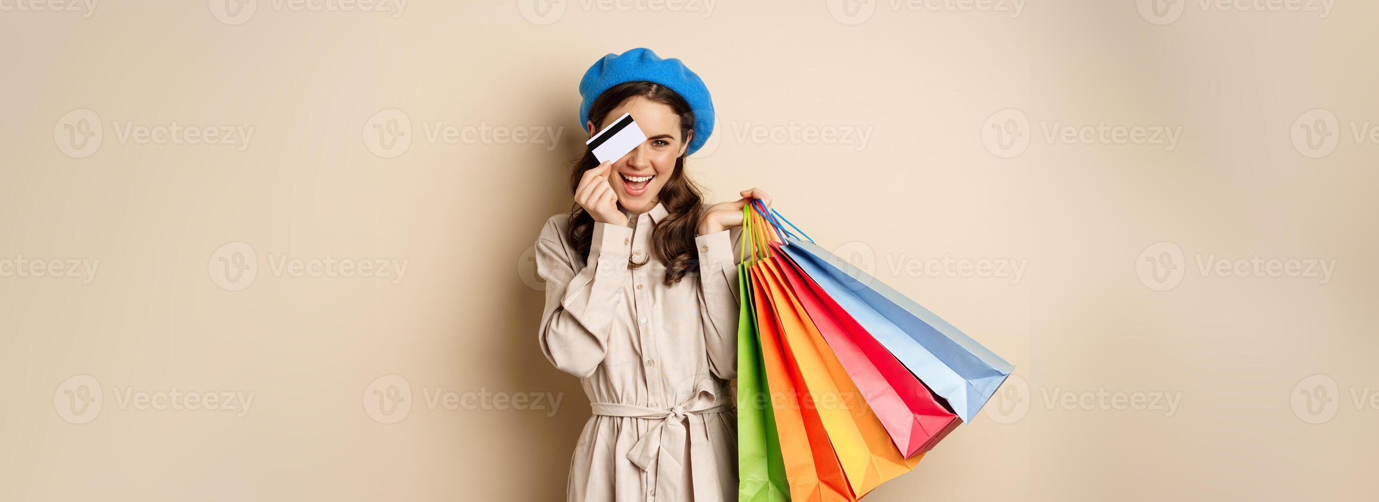 Portrat of trendy feminine girl posing with shopping bags from store and credit card, paying contactless, buying with discount on sale, beige background photo