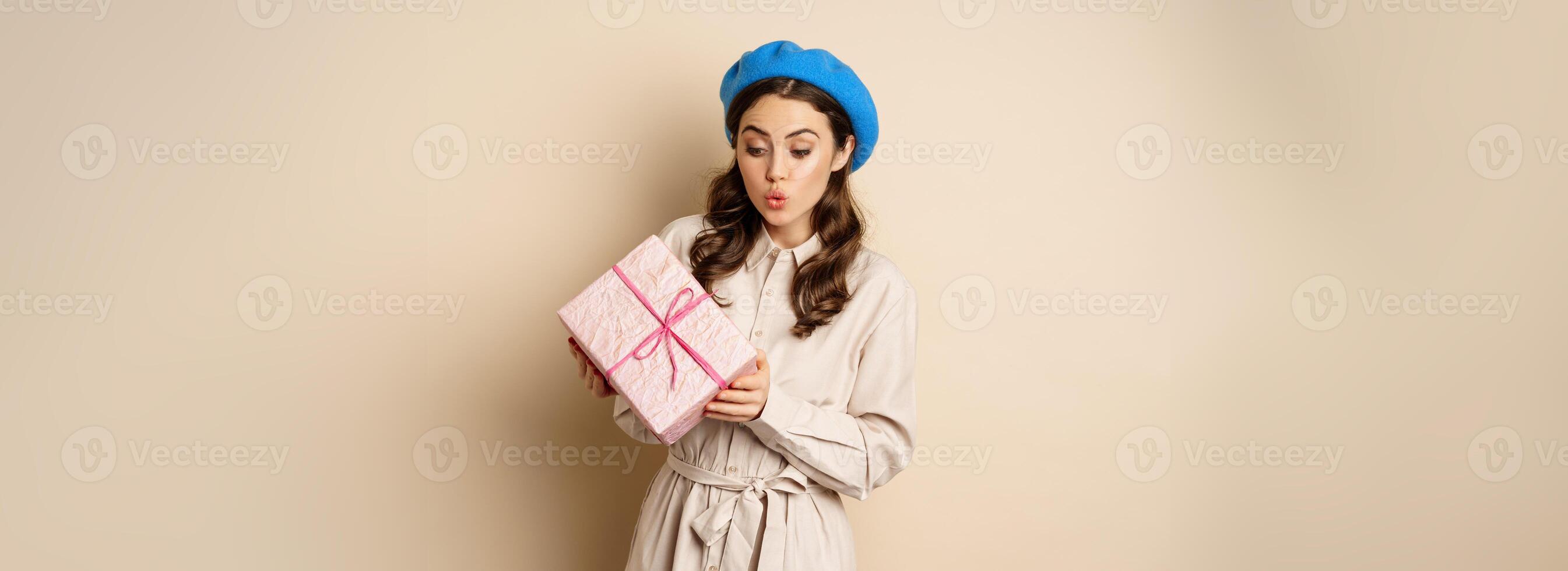 Holidays and gifts concept. Beautiful girl receive gift box and looking happy, holding pink wrapped present with joyful face expression, beige background photo