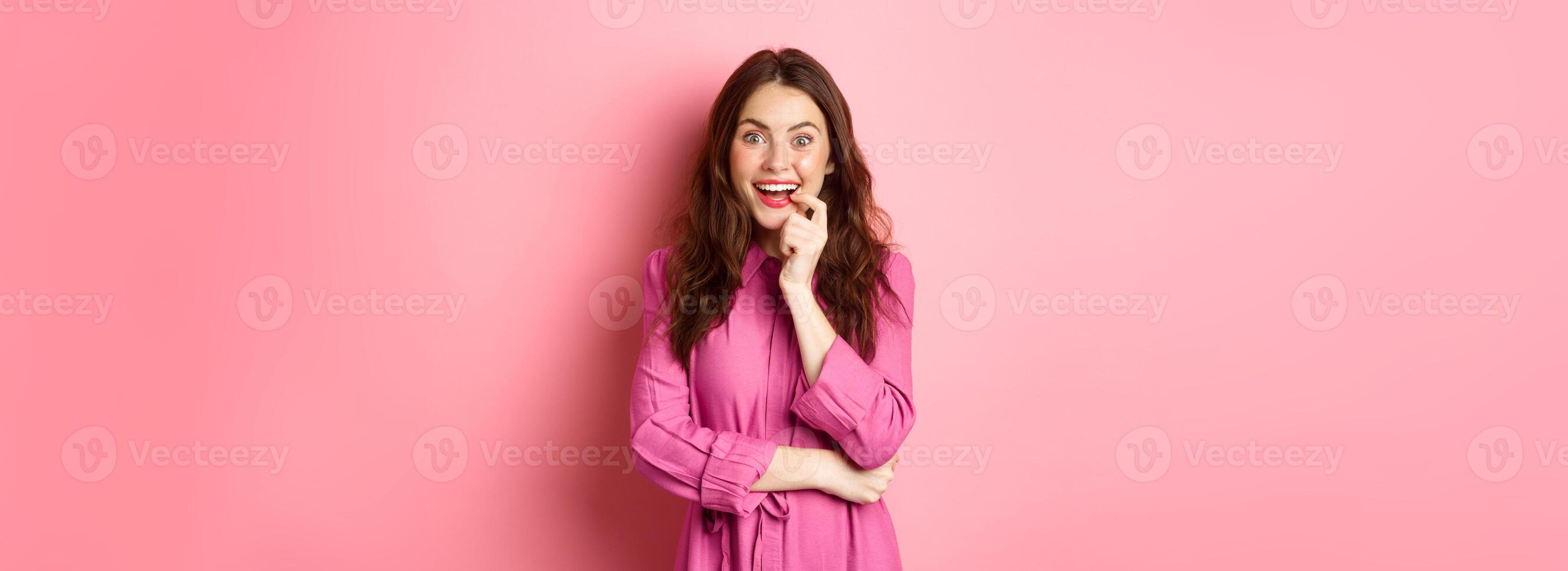 Beautiful excited girl smiling, biting finger and looking with tempted face at camera, having interesting idea, looking at something intriguing, standing against pink background photo