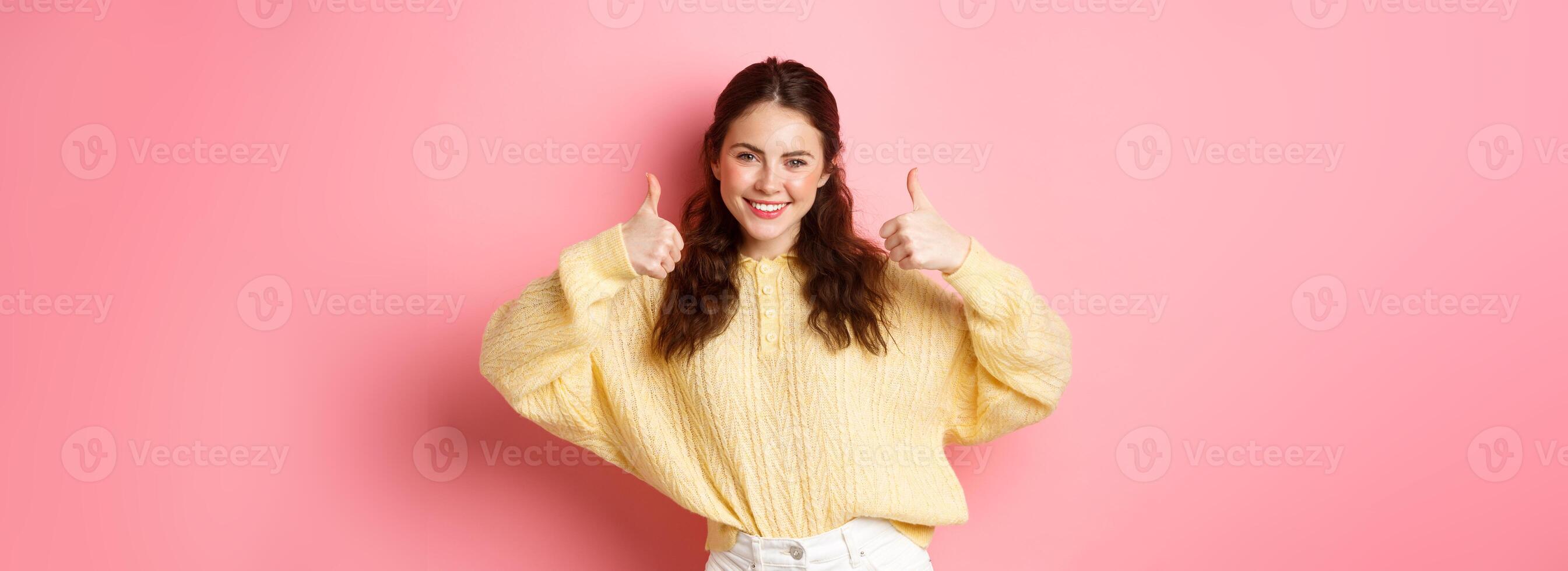 Well done, very good. Smiling young woman showing thumbs up in approval, like your choice, praise good thing, standing satisfied against pink background photo