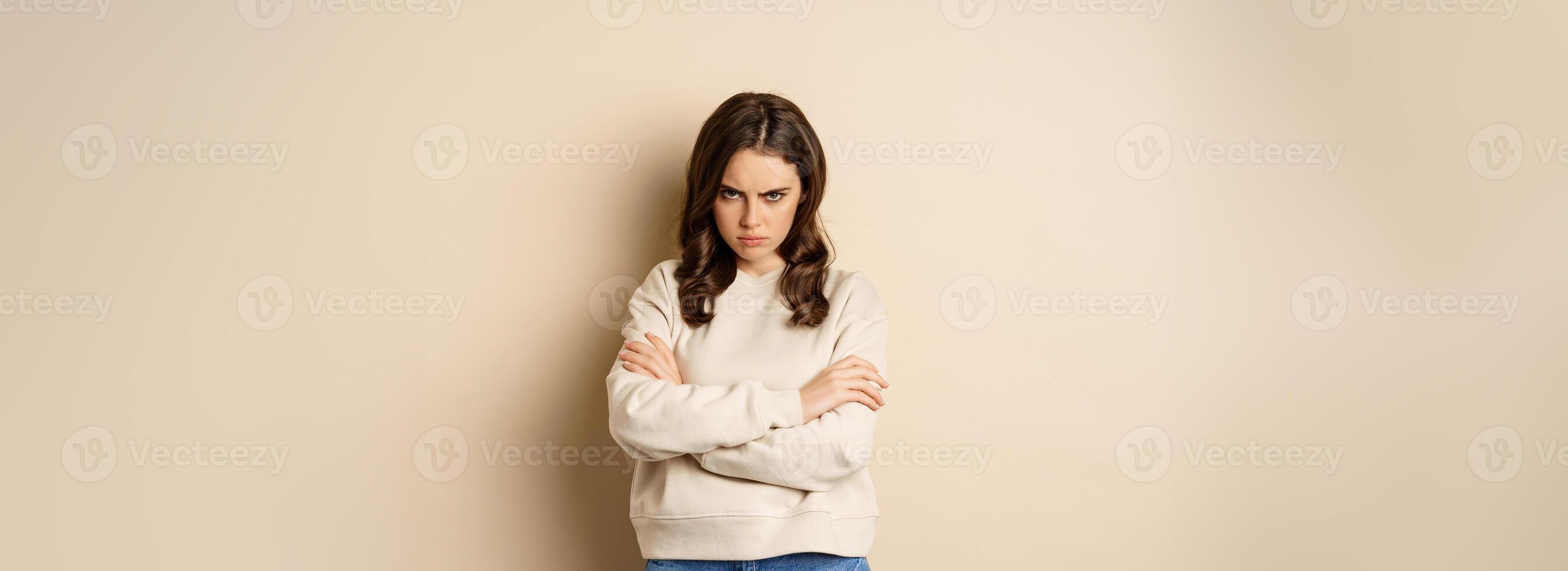 Angry girlfriend sulking, cross arms on chest and look offended, staring moody at camera, standing over beige background photo