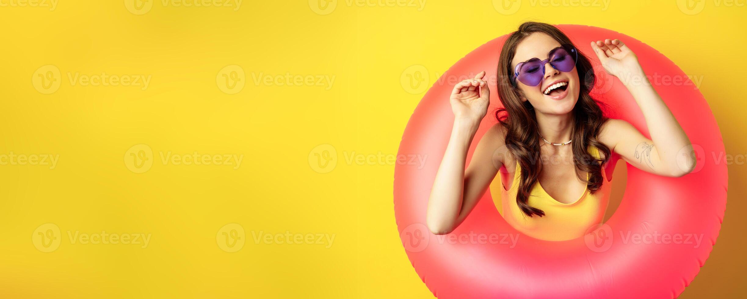 Beautiful happy woman enjoying summer vacation, wearing pink swimming ring on beach, laughing and smiling, standing over yellow background photo
