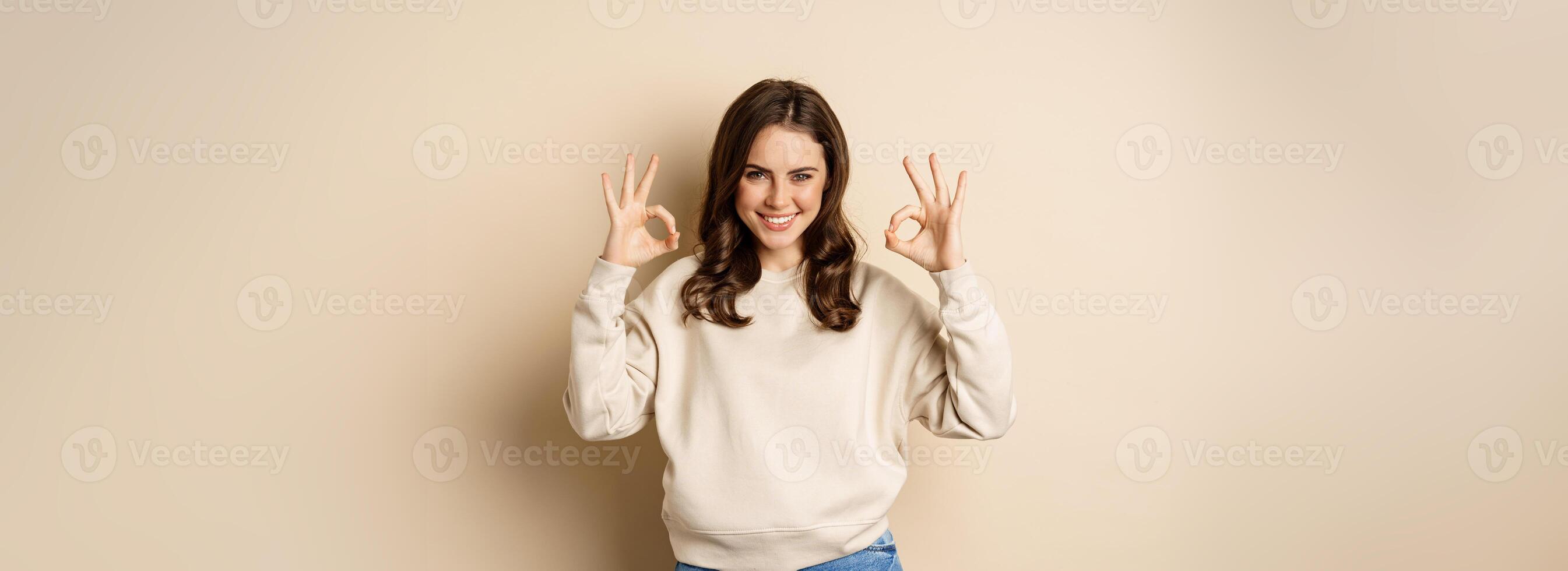 Okay. Confident smiling modern girl showing ok sign, approve, like and recommend smth good, standing over beige background photo