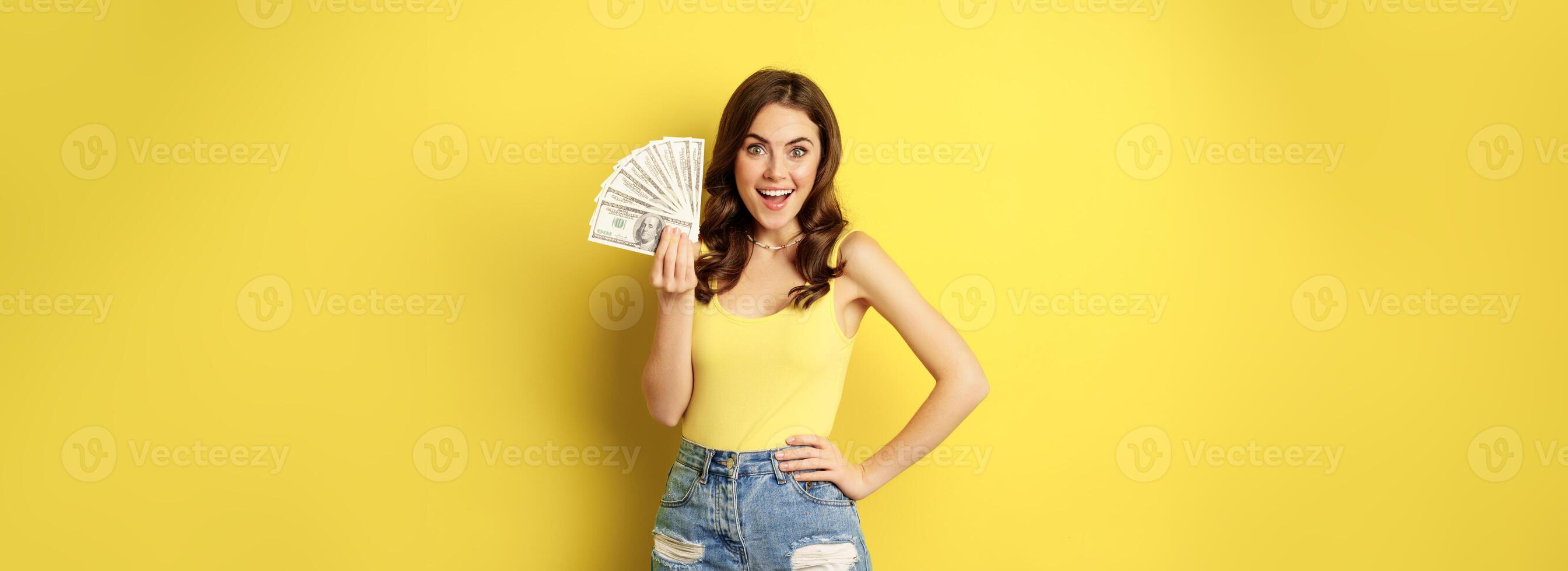 Shopping, credit and money concept. Young brunette woman showing cash and smiling pleased, standing over yellow background photo