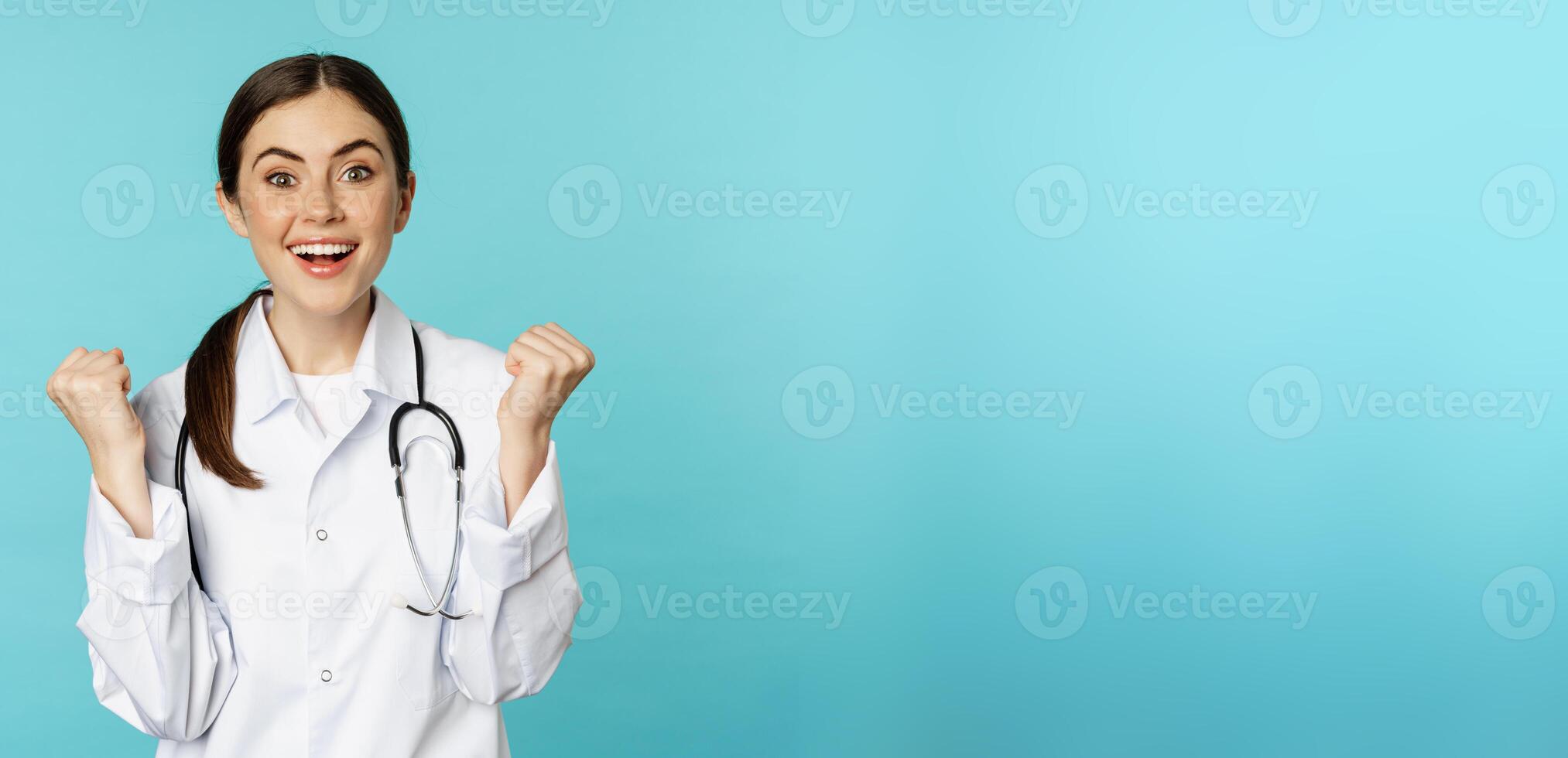 Happy and excited young female doctor, physician celebrating, achieve goal and smiling pleased, winning, standing in white coat against blue background photo