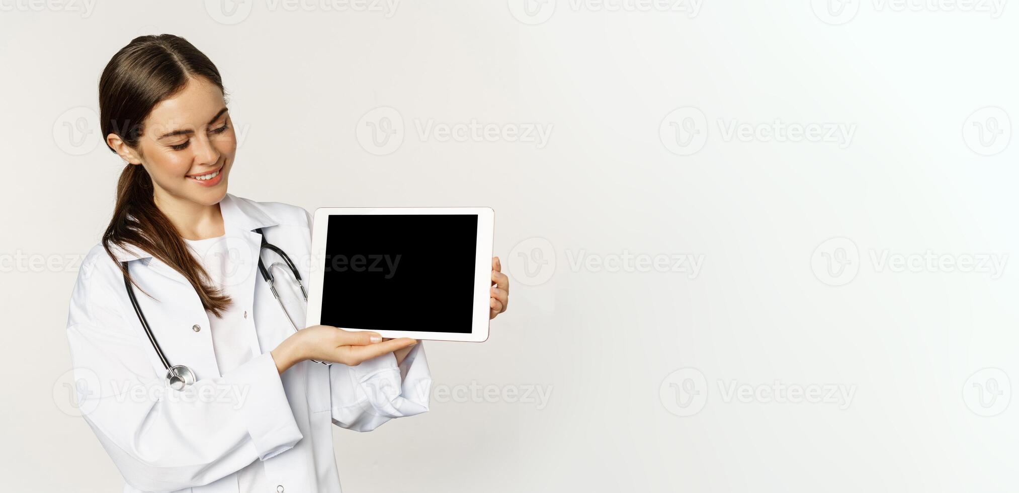 Smiling doctor, female physician or nurse showing digital tablet screen, demonstrating online website, application, standing over white background photo