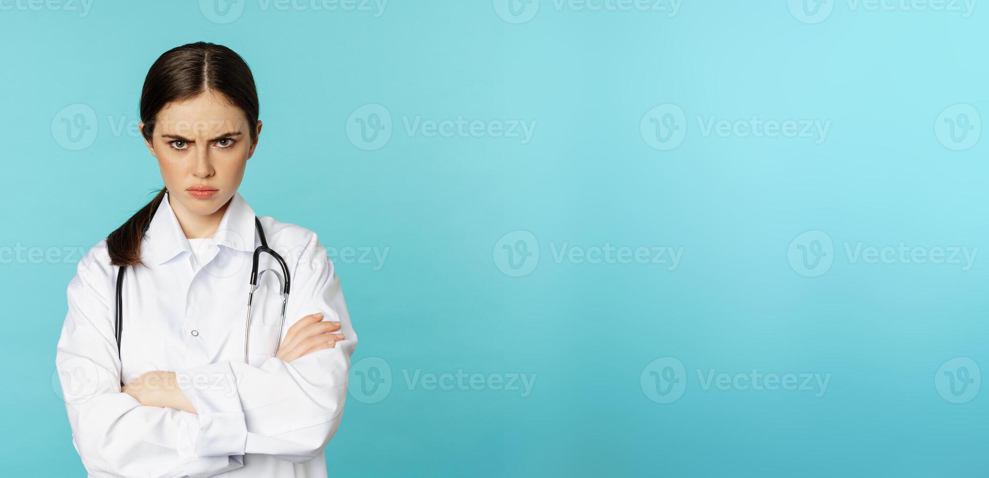 Angry doctor, female medical worker looking offended, cross arms on chest, frowning and staring annoyed at camera, standing over blue background photo