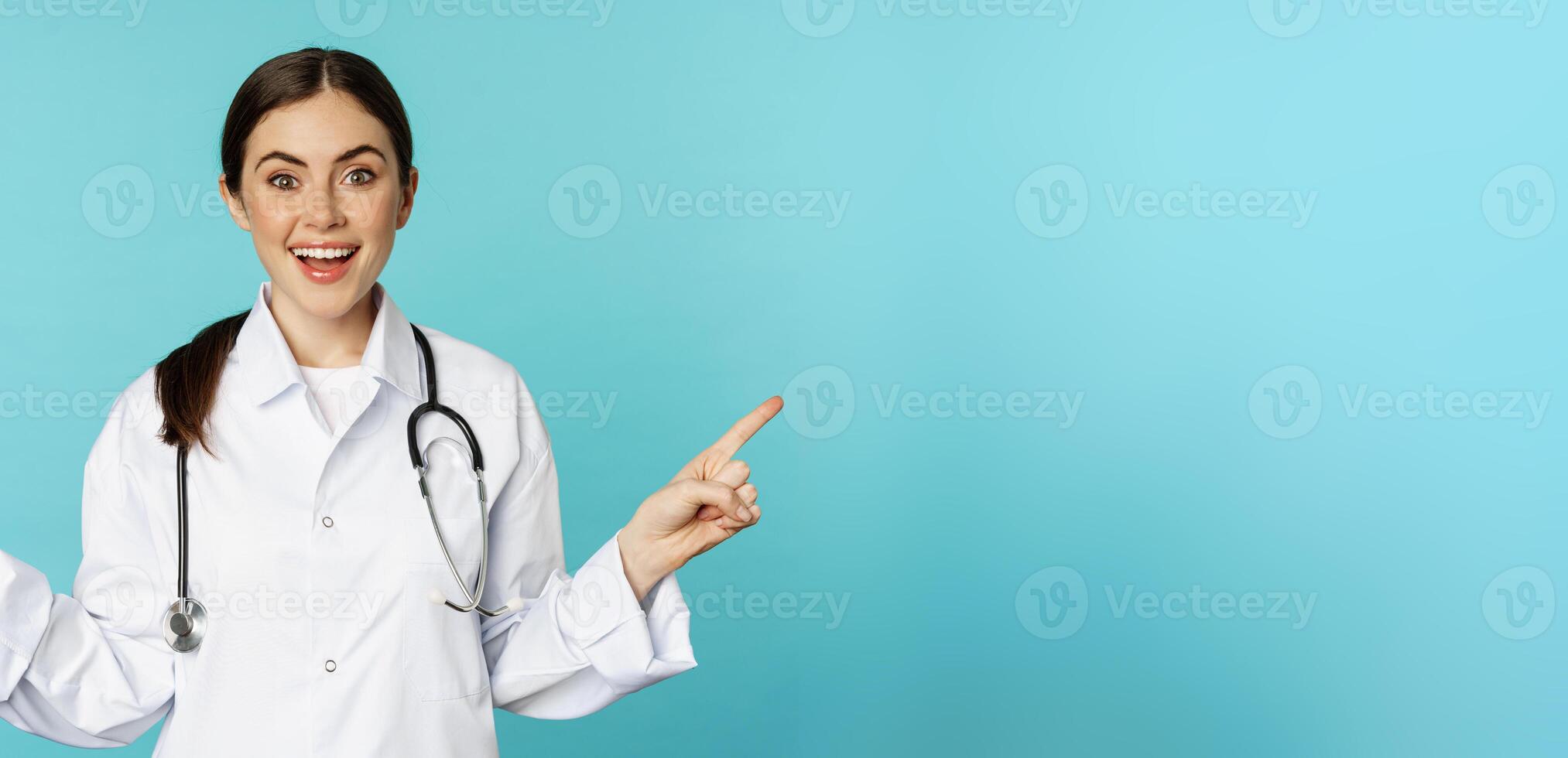 Enthusiastic woman doctor, medical worker in white coat, pointing fingers sidweays, left and right, showing health clinic promo, standing over torquoise background photo