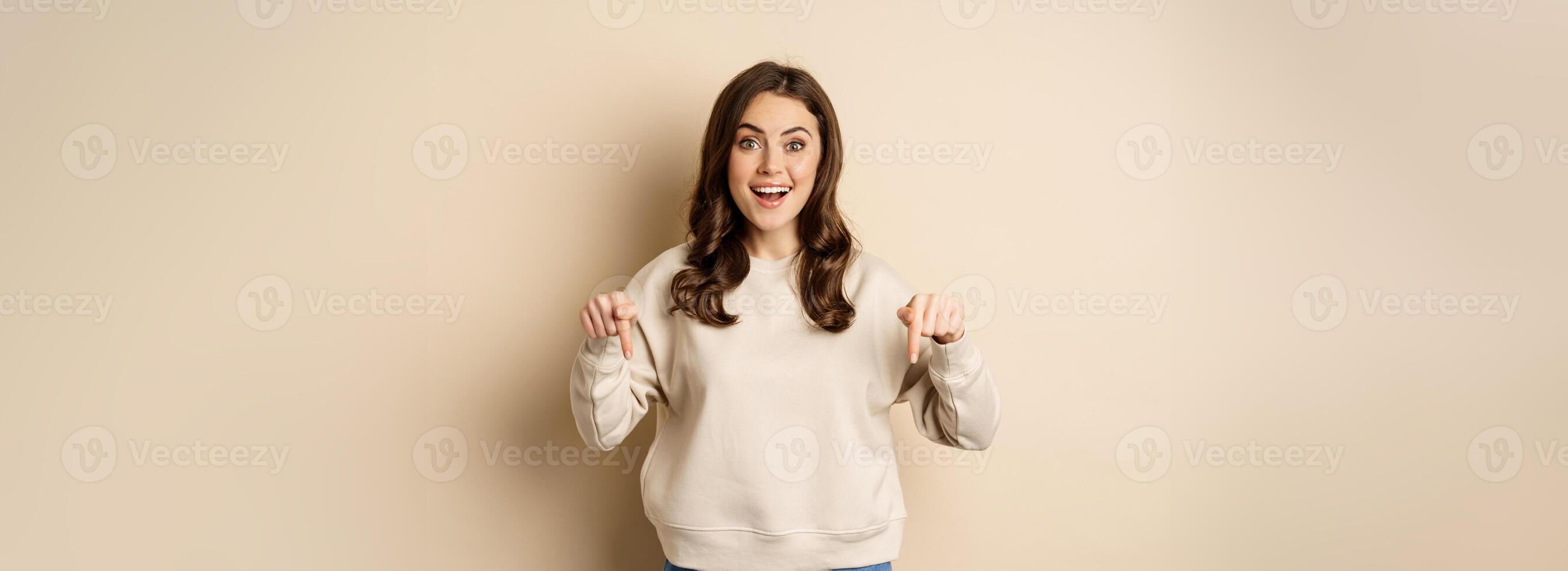 Enthusiastic attractive girl pointing fingers down, showing advertisement, link or promo below, standing over beige background photo