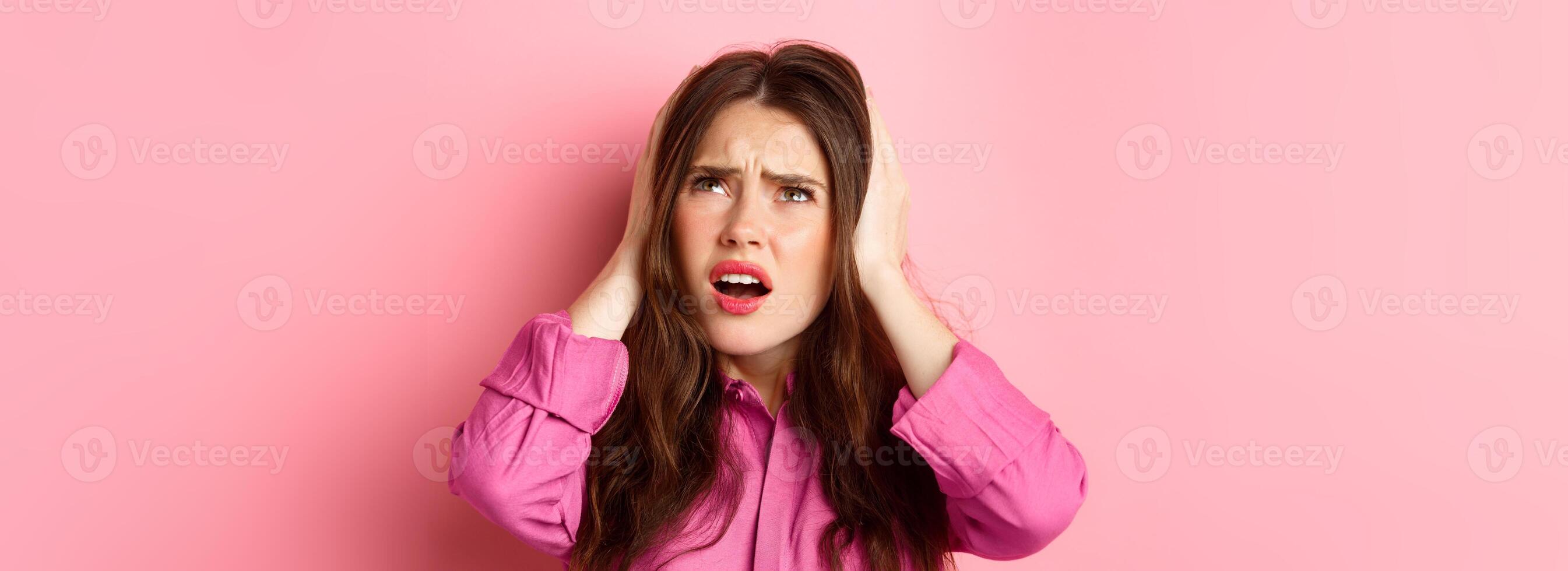 Close up of annoyed glamour girl covering ears with hands, looking bothered up at neighbours, cant stand loud music, standing over pink background photo