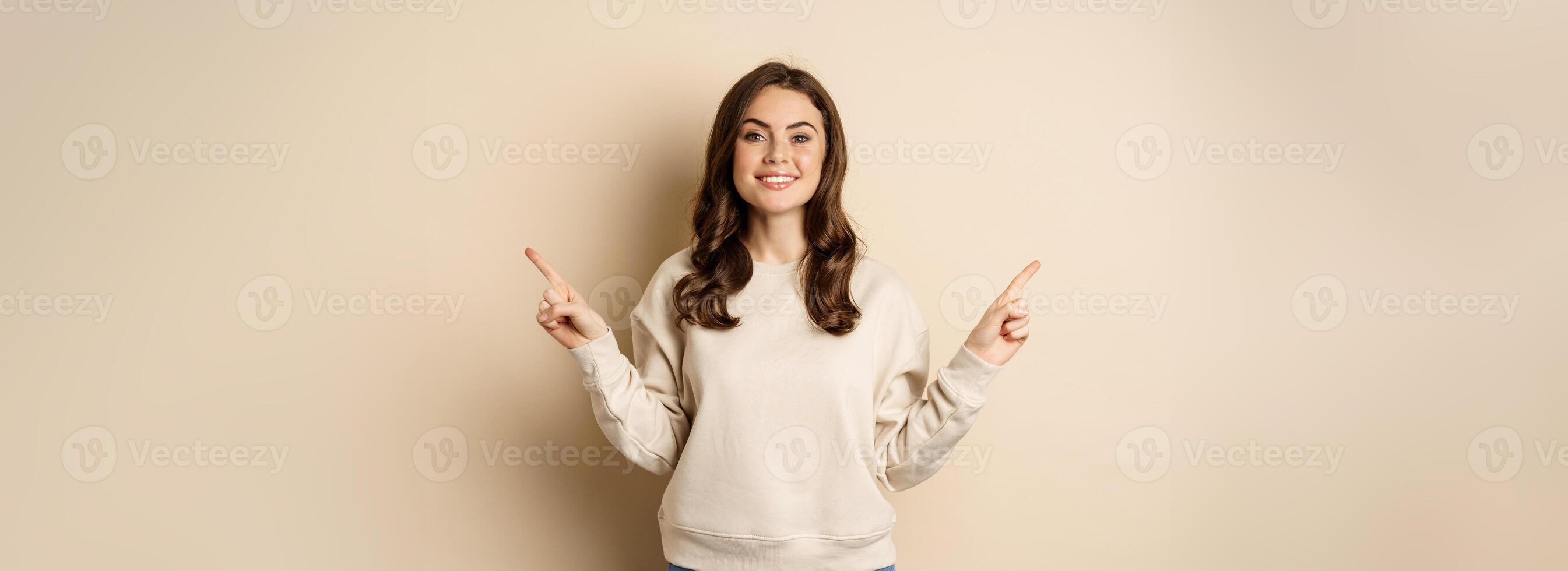 Cheerful caucasian woman showing directions, two ways, pointing sideways at variants, choices for customer, standing over beige background photo