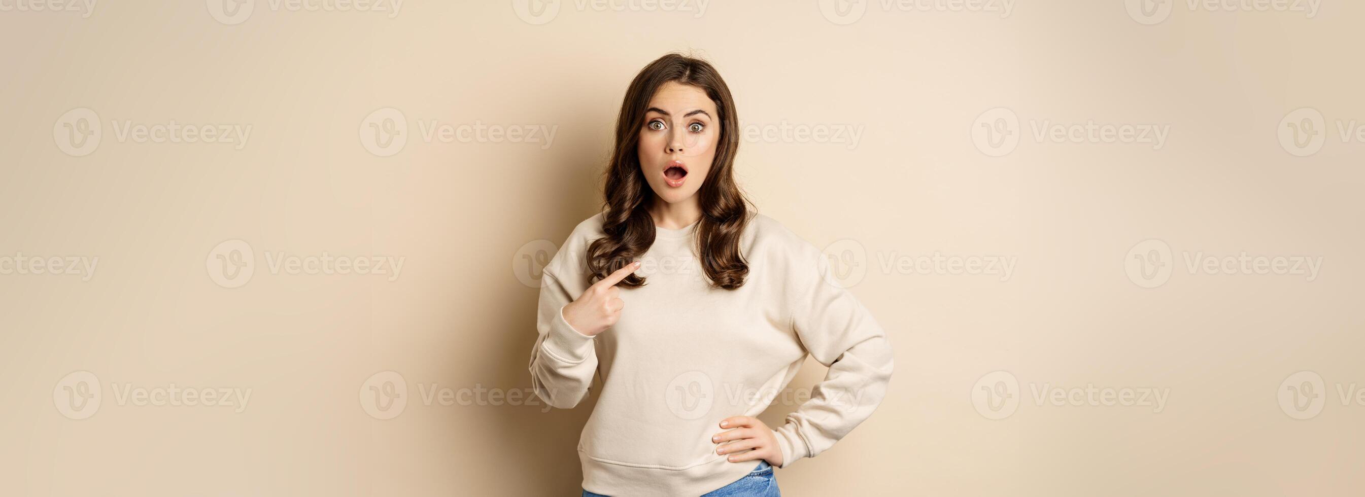 Surprised girl pointing at herself and look with disbelief, standing over beige background photo