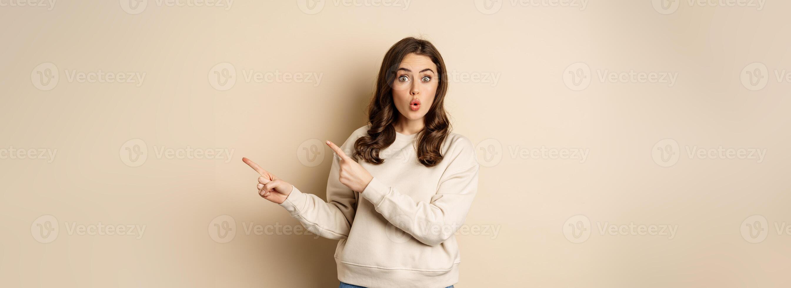 Surprised brunette woman pointing fingers right, showing advertisement and say wow impressed, standing over beige background photo