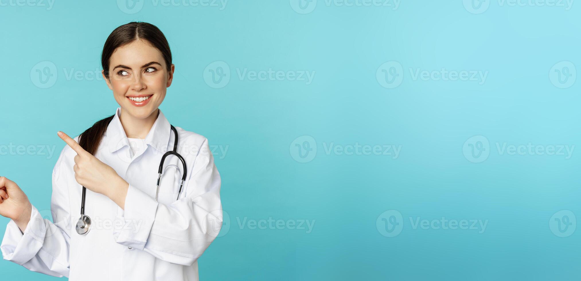 Portrait of smiling medical worker, girl doctor in white coat with stethoscope, pointing fingers left, showing medical clinic advertisement, torquoise background photo