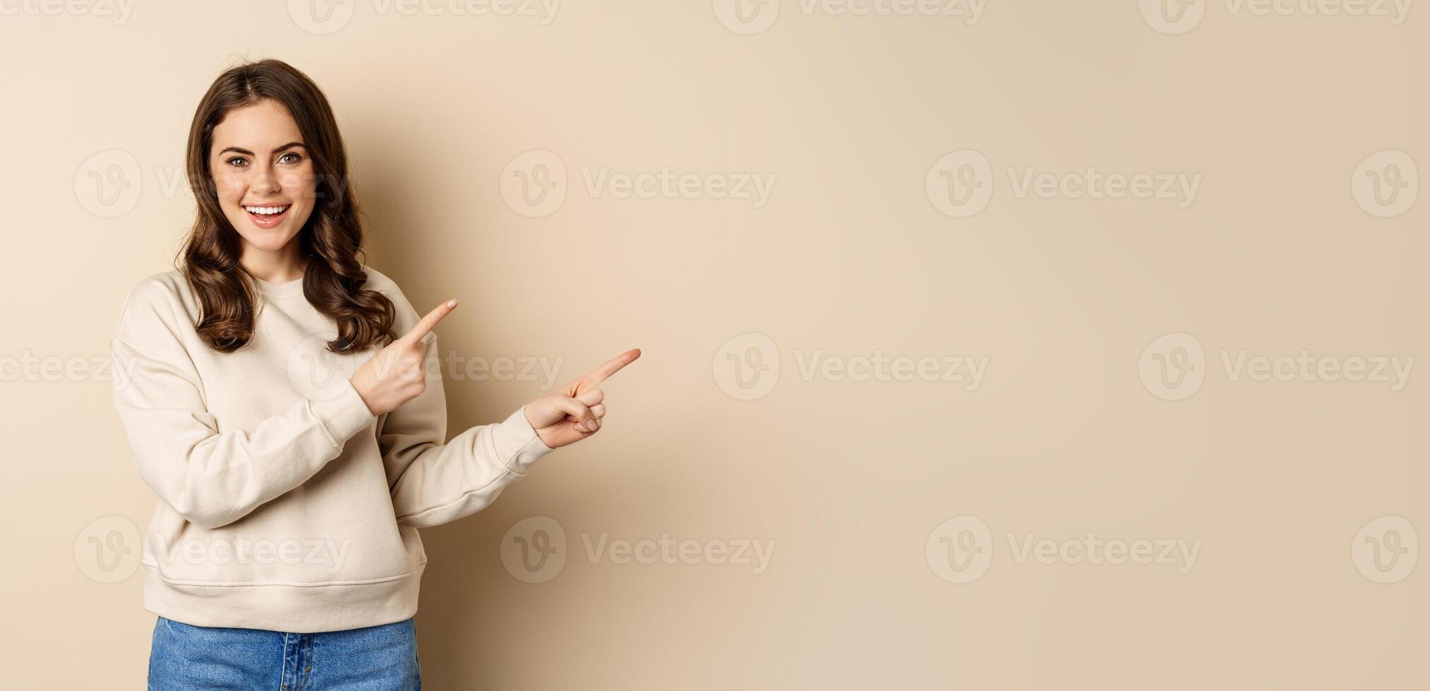 Smiling gorgeous woman pointing fingers right, inviting people, showing logo or banner, standing over beige background photo