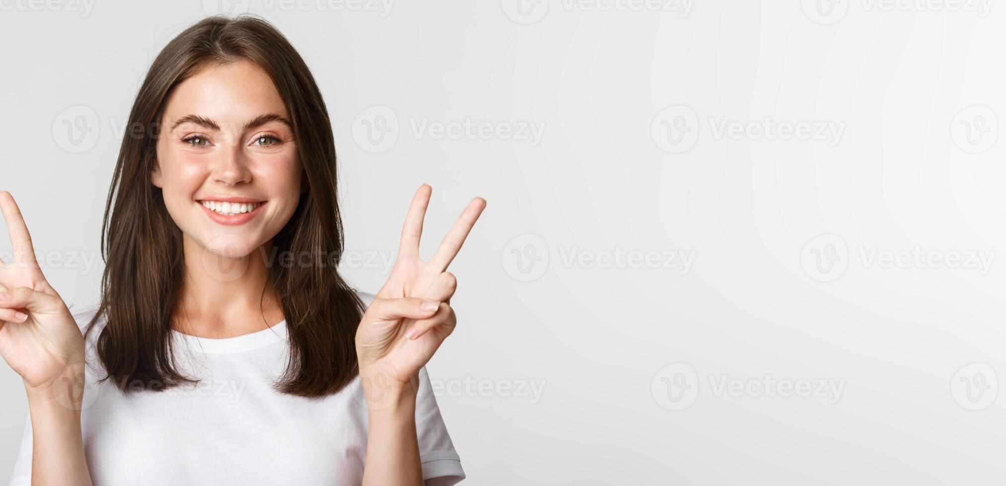 Portrait of beautiful and happy brunette girl smiling and showing peace gestures, standing white background photo