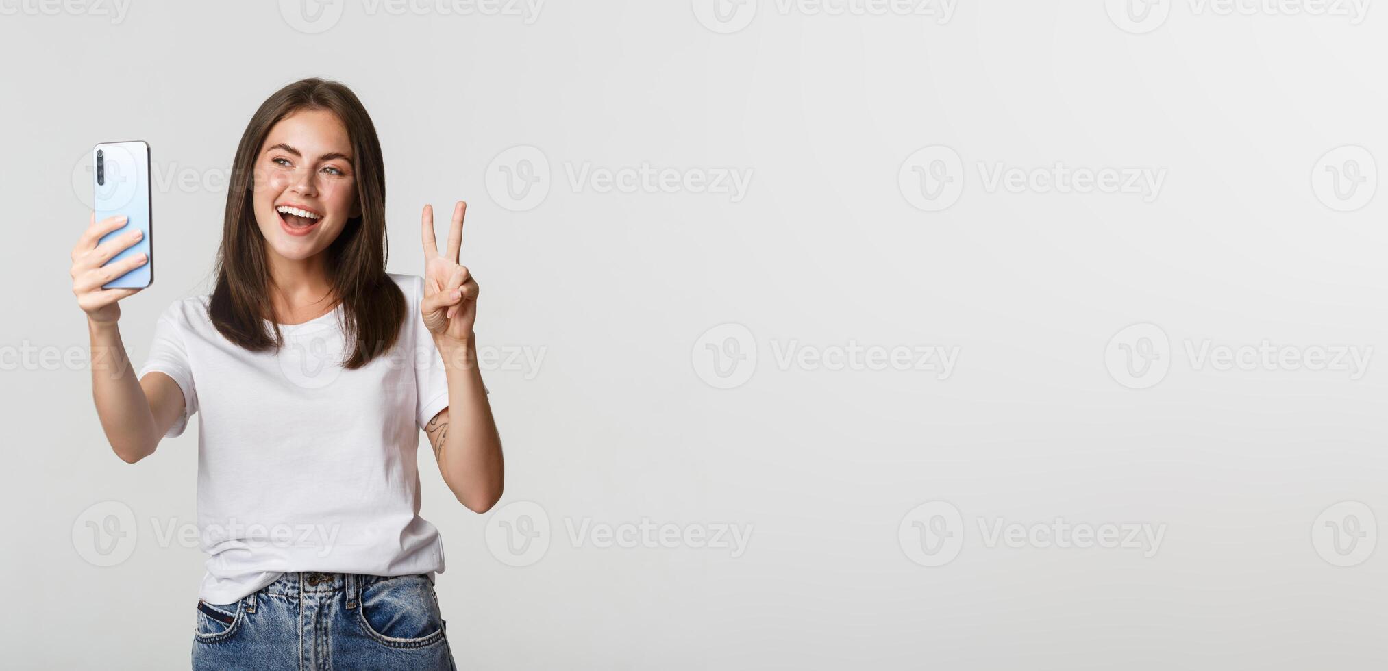 Attractive brunette girl looking happy and taking selfie on smartphone, showing peace gesture photo