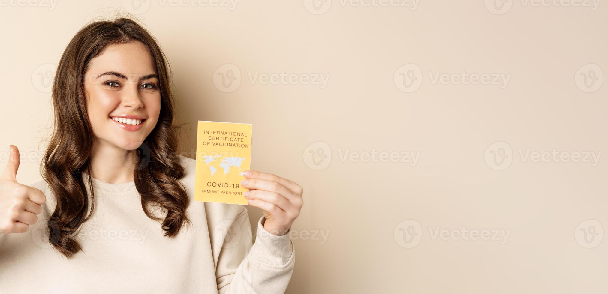 Covid-19 vaccination concept. Portrait of happy healthy girl showing coronavirus international vaccination certificate and thumb up, travelling during pandemic, beige background photo