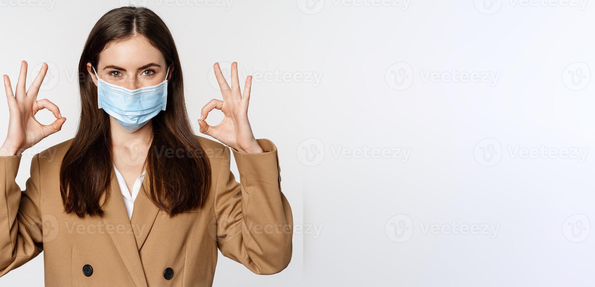 Workaplce and pandemic concept. Close up portrait of business woman showing okay signs, wearing medical mask, recommend, praise something, standing over white background photo