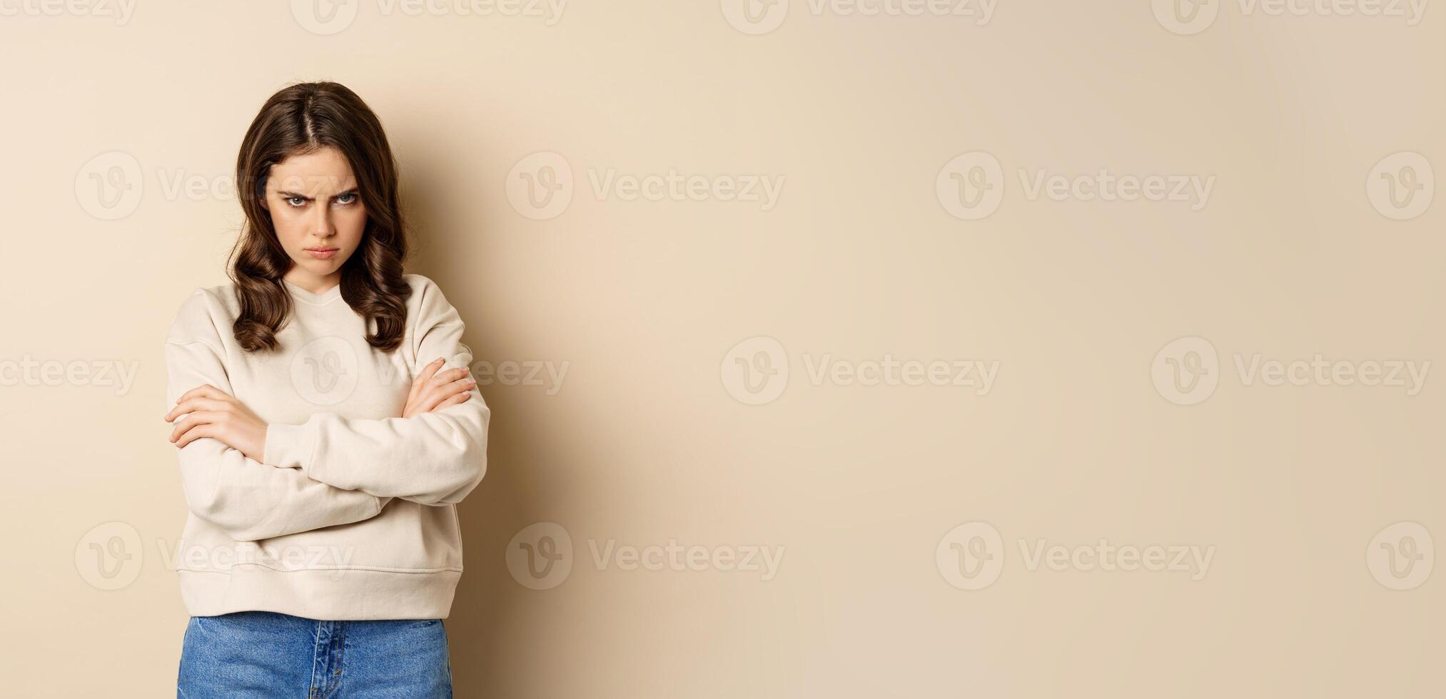 Angry girlfriend sulking, cross arms on chest and look offended, staring moody at camera, standing over beige background photo