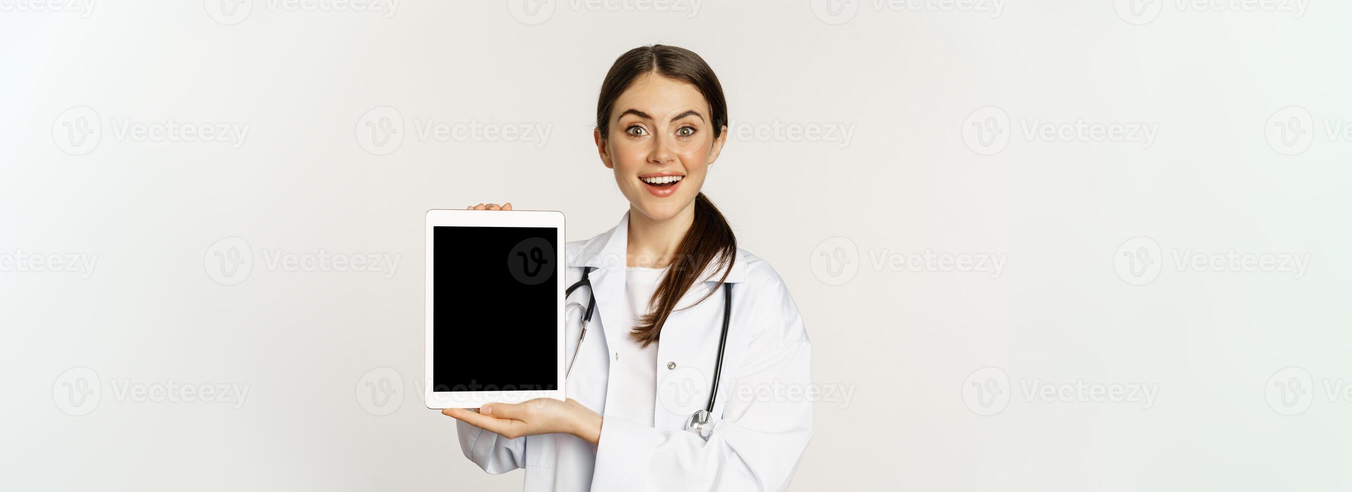 Image of woman doctor, female healthcare worker showing online medical website, digital tablet screen and smiling, standing in white coat over white background photo