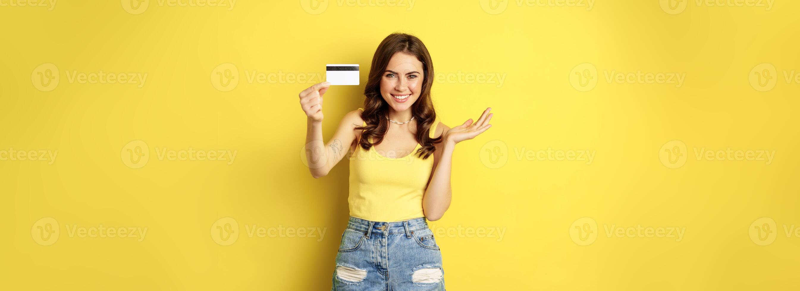 Portrait of brunette girl showing her credit card and smiling, wearing summer clothes, concept of bank, money and shopping, standing over yellow background photo