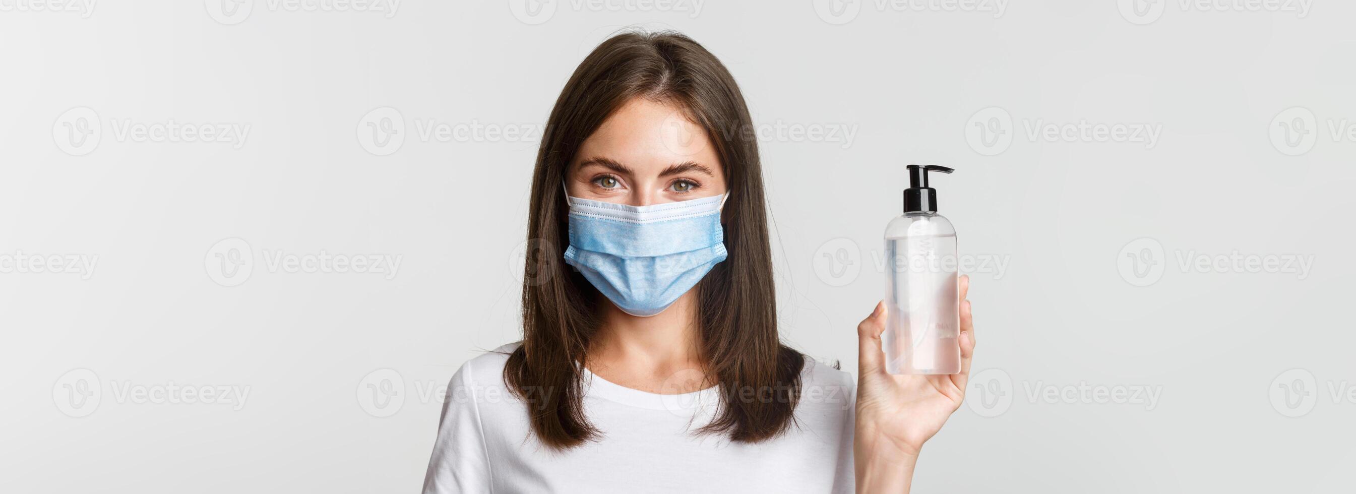Covid-19, health and social distancing concept. Close-up of attractive smiling girl in medical mask, showing hand sanitizer, recommend antiseptic photo