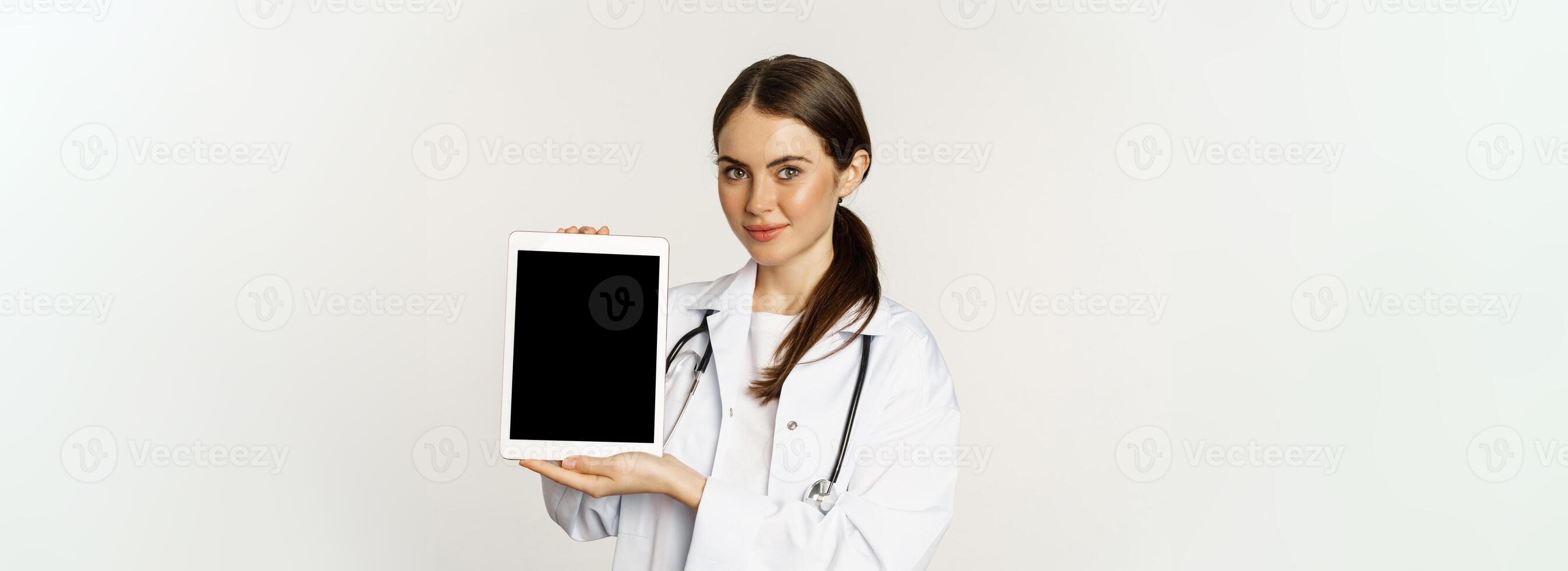 Image of woman doctor, female healthcare worker showing online medical website, digital tablet screen and smiling, standing in white coat over white background photo