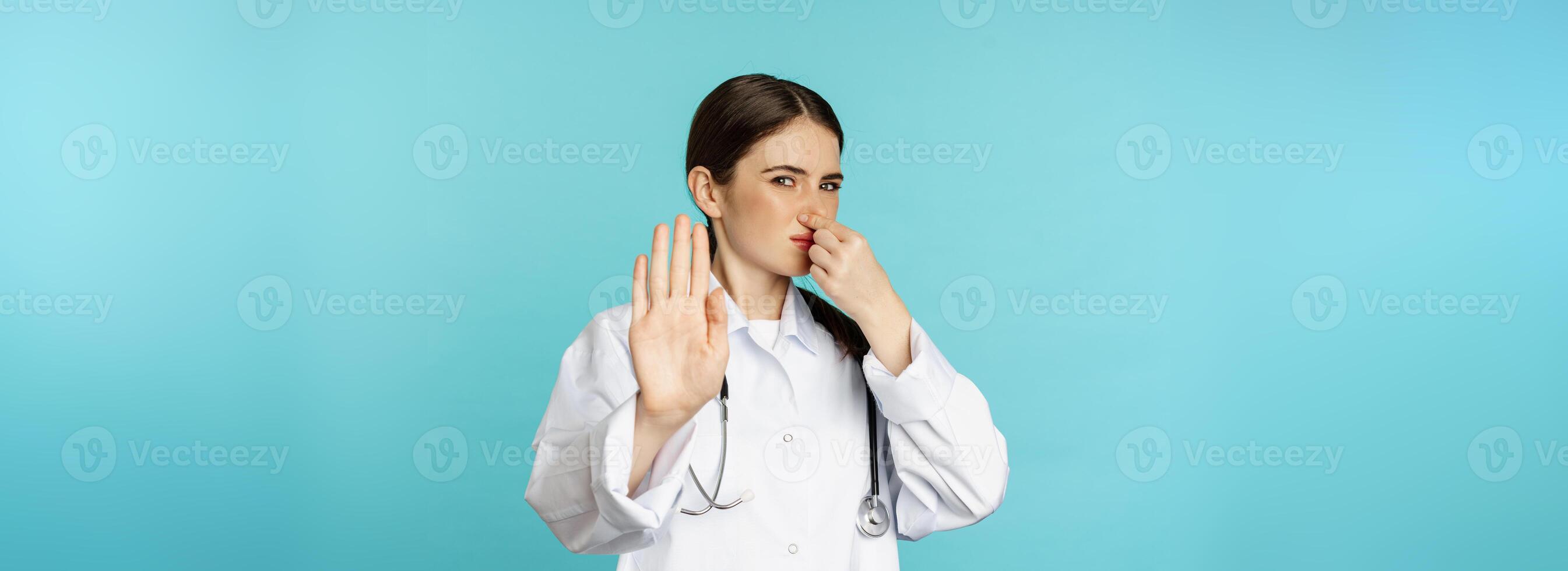 Woman doctor dislike, shut nose from bad smell, reek, showing rejection, stop gesture, standing against torquoise background photo
