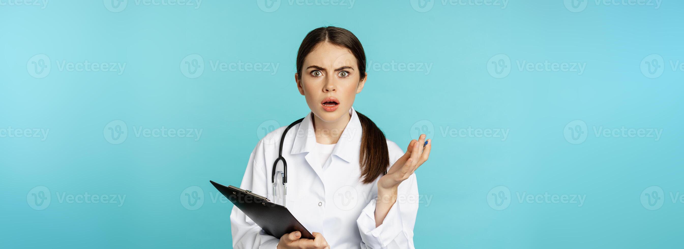 Shocked woman doctor reacting worried and frustrated at camera, hear smth strange, standing in white coat against torquoise background photo