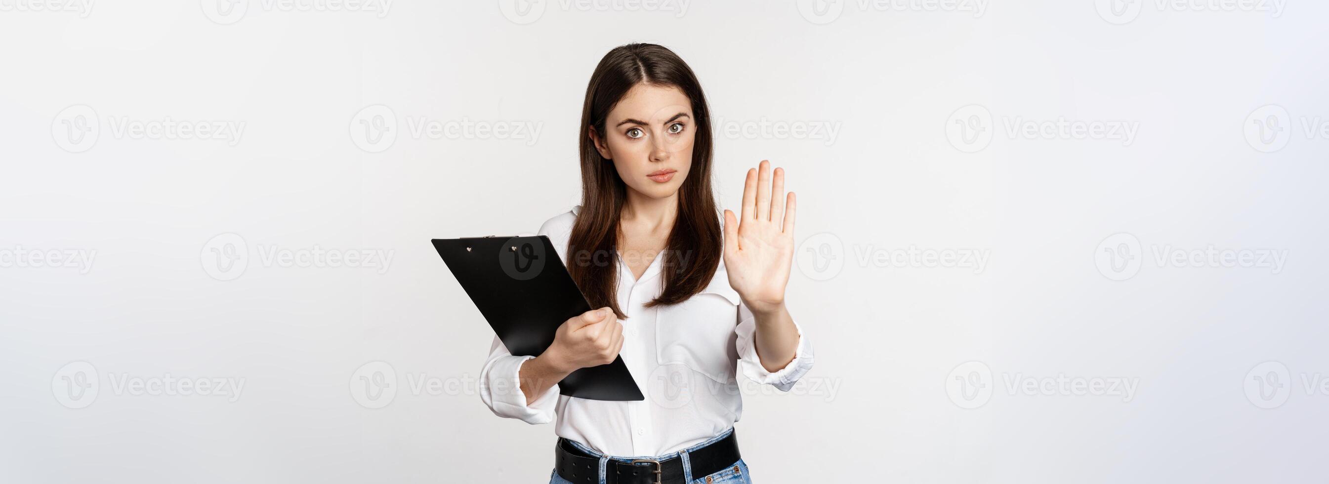 Serious woman with clipboard documents, extending arm, showing taboo stop sign, disapprove, prohibit smth, standing over white background photo
