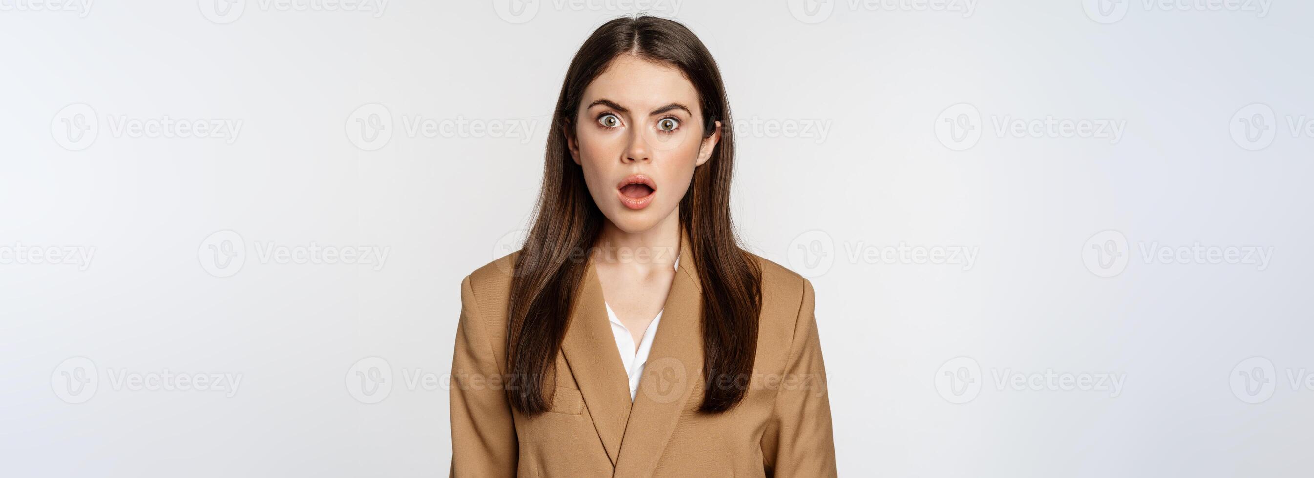 Portrait of shocked, speechless businesswoman gasping, staring stunned at camera, wearing brown suit, white background photo