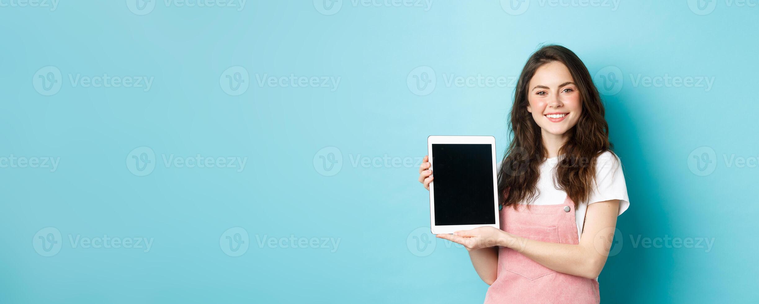 Portrait of smiling beautiful woman demonstrate blank tablet screen, showing promo on device screen, standing against blue background photo