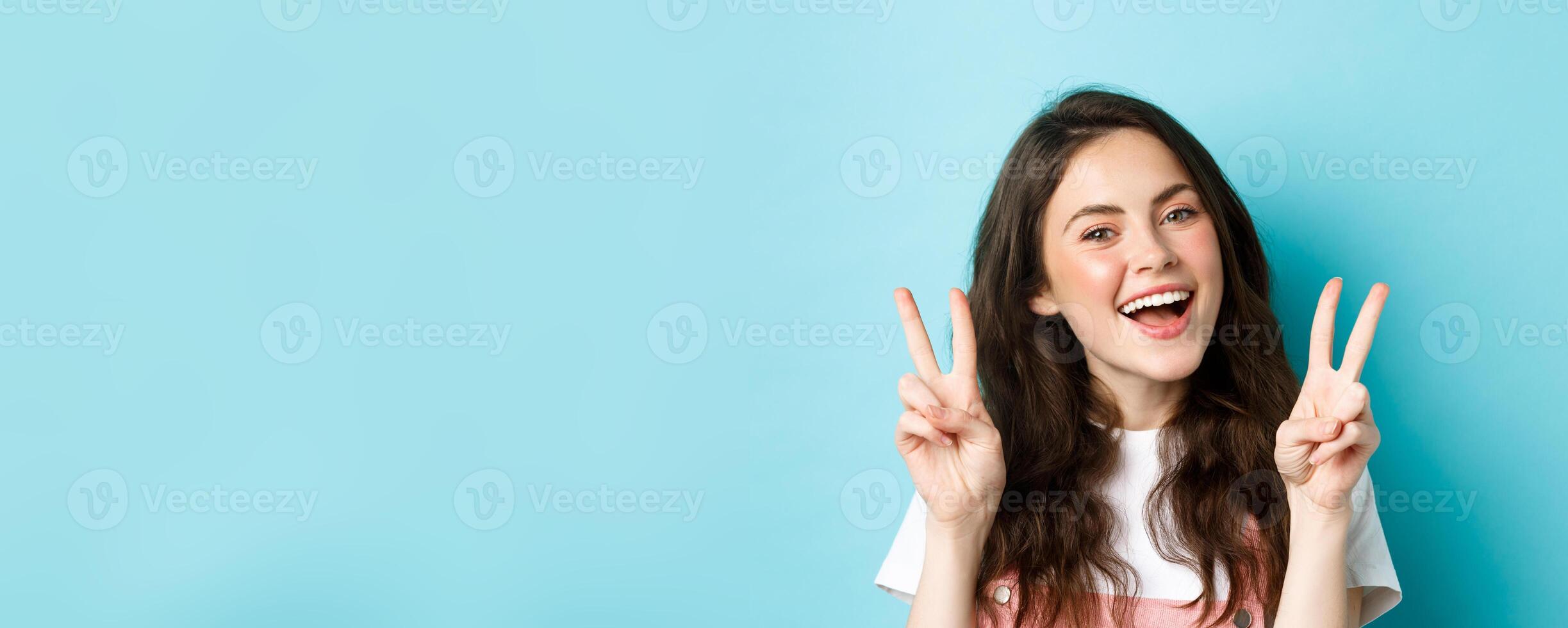 Young positive modern girl sending good vibes only, showing v-sign and smiling happy, standing against blue background photo