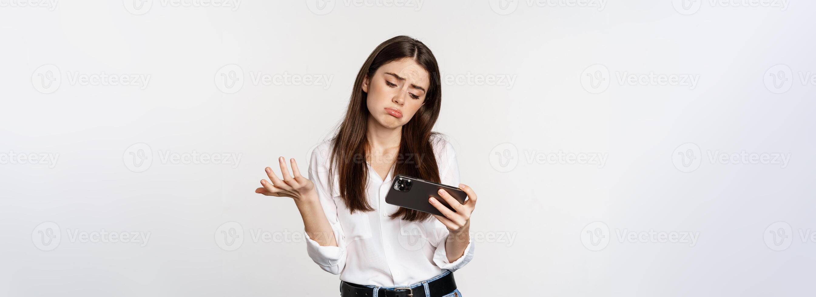 Sad woman losing in mobile game, looking upset and disappointed at smartphone, sulking, standing over white background photo