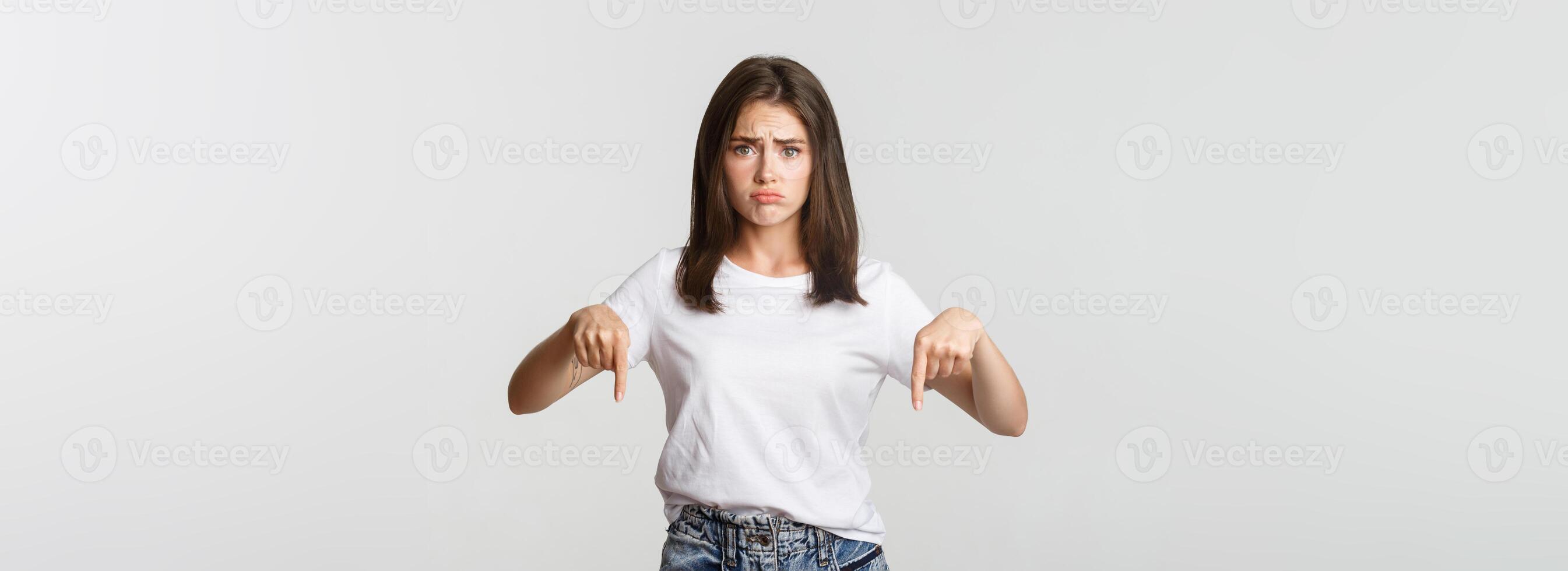 Sad and gloomy brunette girl sulking, pointing fingers down with regret photo