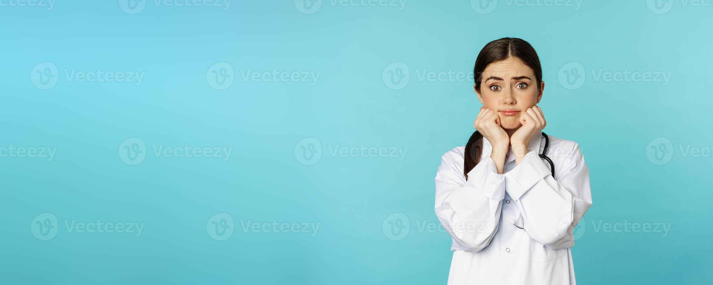 Scared and worried woman doctor in white coat, looking anxious and insecure, shaking from fear, standing over torquoise background photo