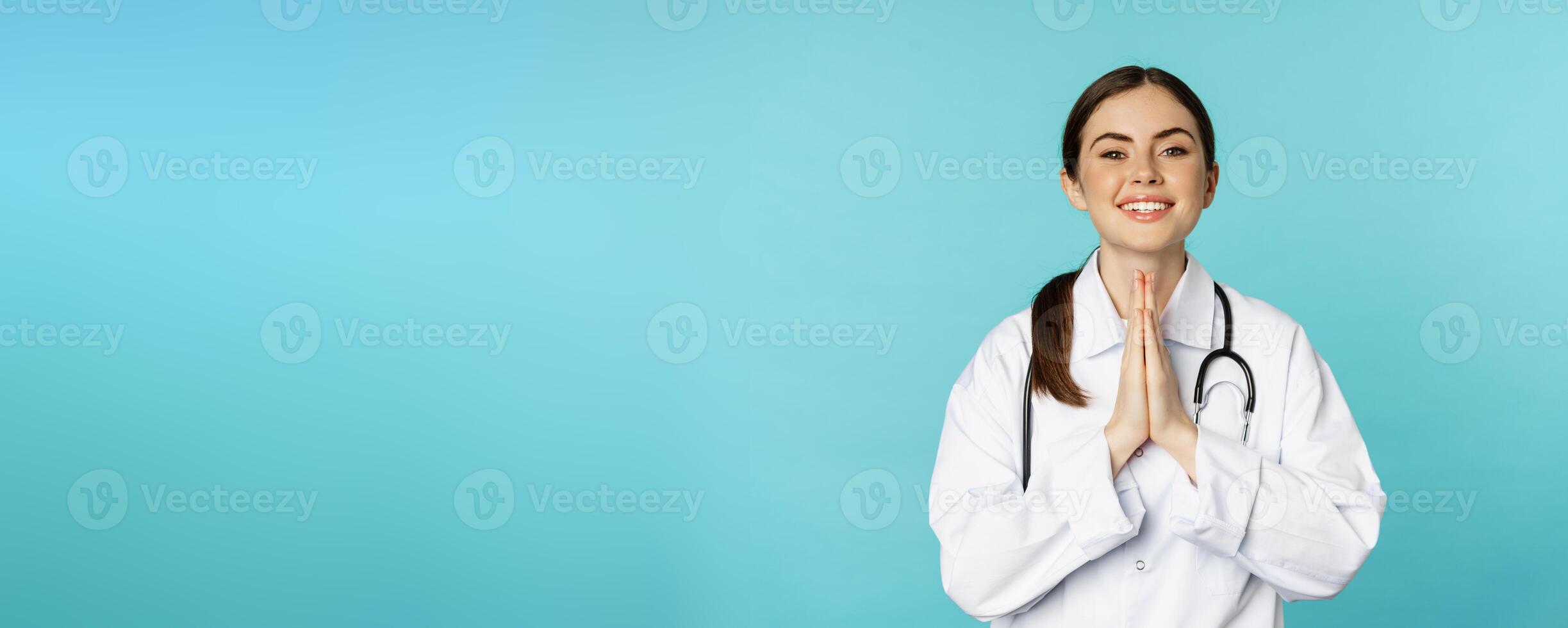 Smiling hopeful young woman doctor, holding hands in begging, thank you pose, standing over blue background photo