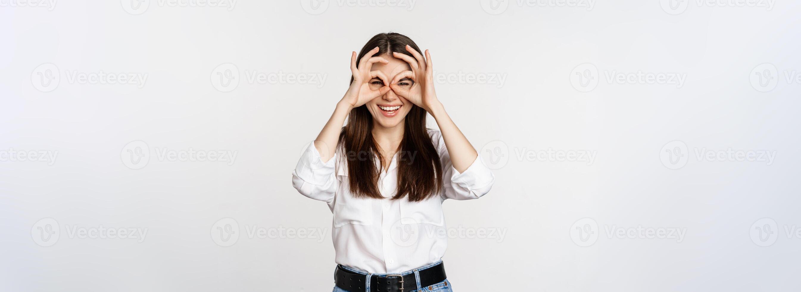 Funny girl looking through hand binoculars, zero gesture, standing over white background and smiling, seeing smth interesting far away, white background photo