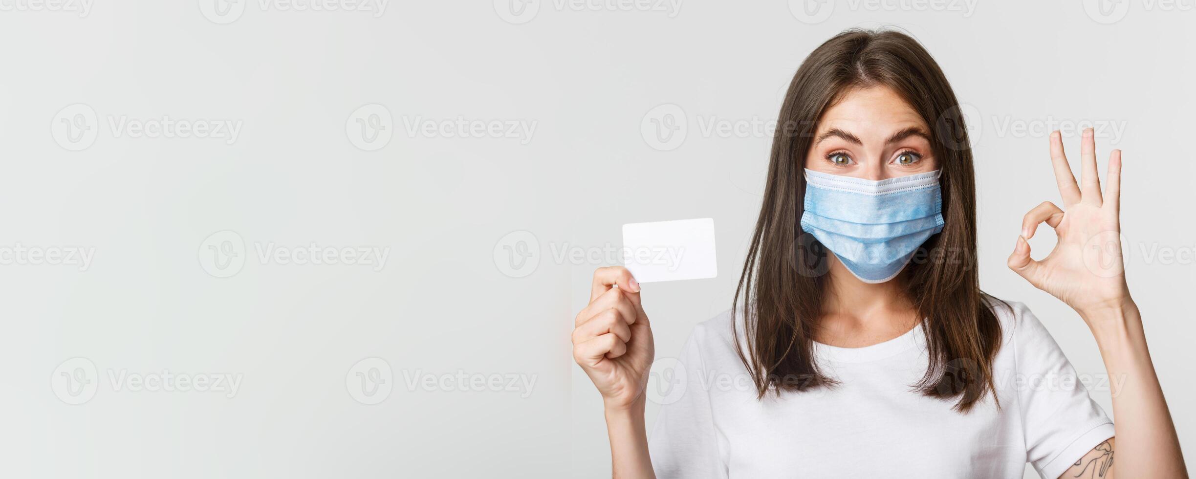 Covid-19, health and social distancing concept. Close-up of smiling brunette girl in medical mask, showing credit card and okay gesture photo