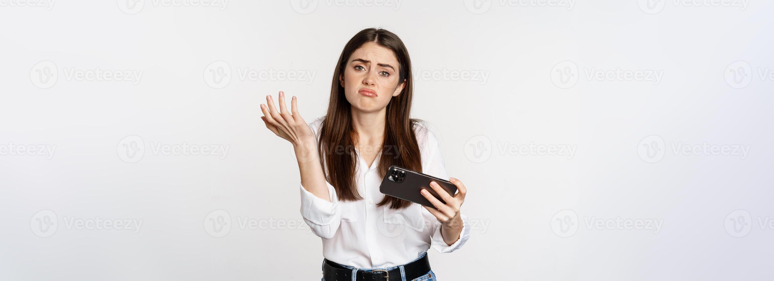 Sad woman losing in mobile game, looking upset and disappointed at smartphone, sulking, standing over white background photo