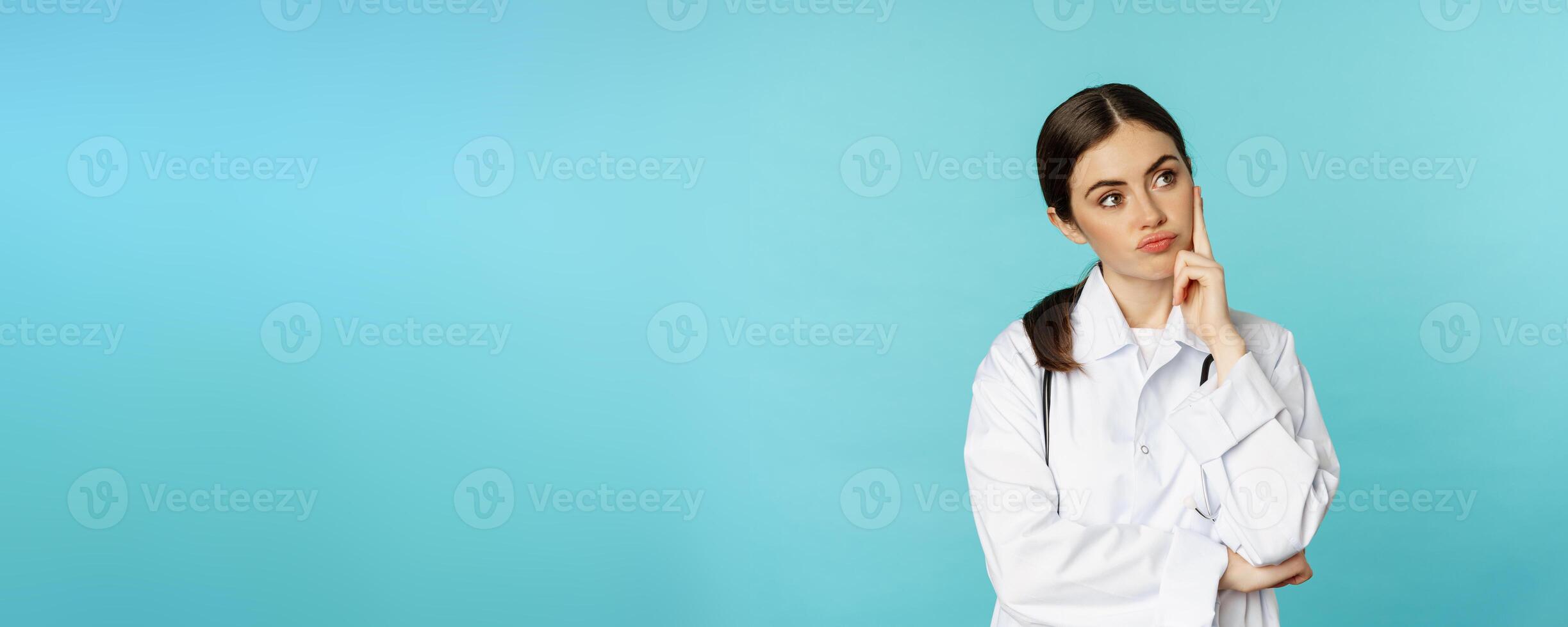 Thoughtful medical worker, young doctor woman looking unamused, bored, standing in white coat against blue background photo